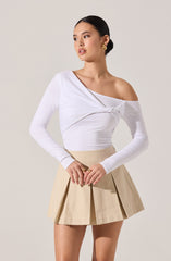 A woman is wearing a white, long-sleeved, off-the-shoulder top with a gathered detail across the front. She pairs it with a beige, pleated, high-waisted mini skirt.