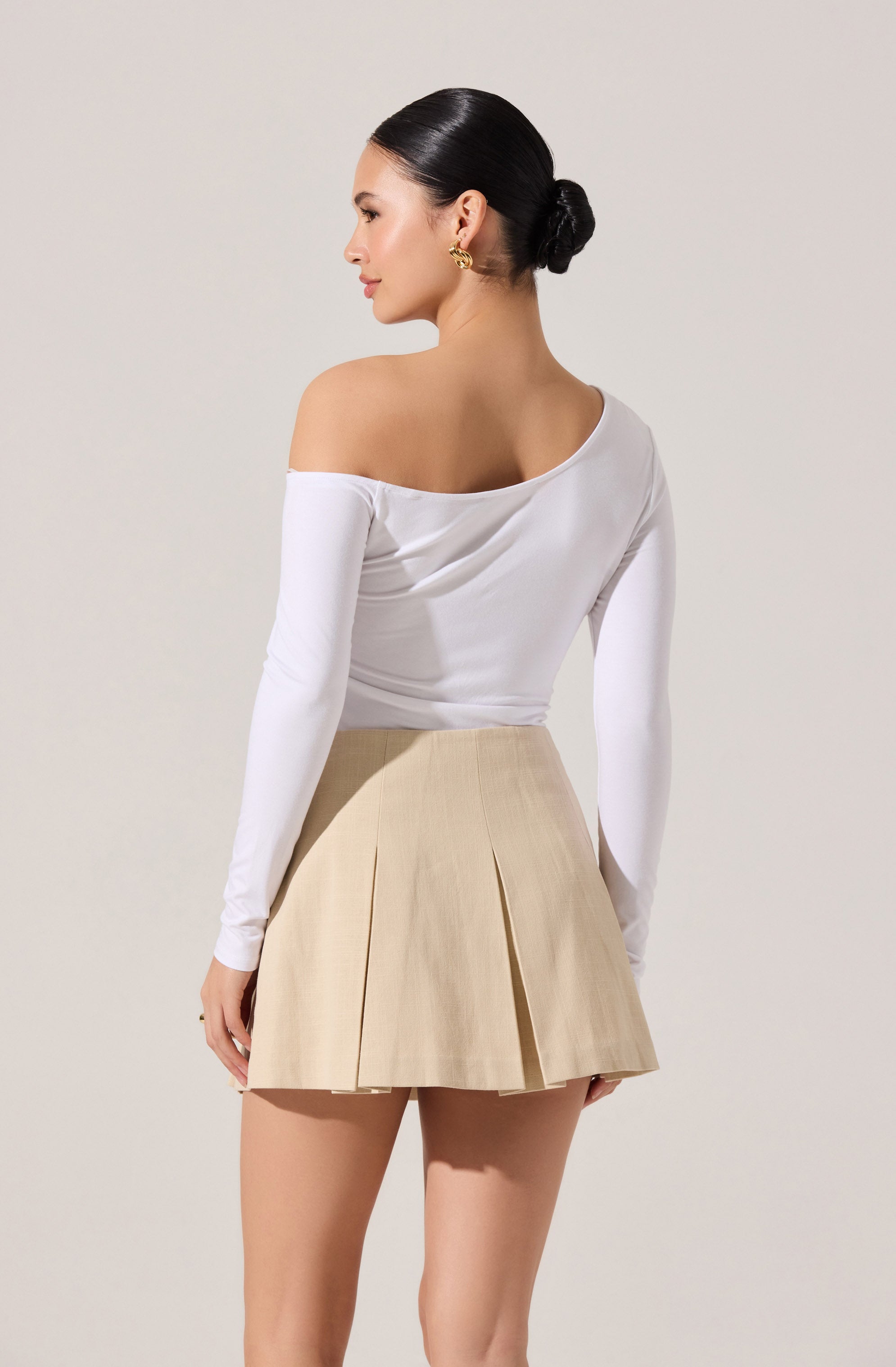 A woman is wearing a white off-the-shoulder long-sleeve top and a beige pleated mini skirt.
