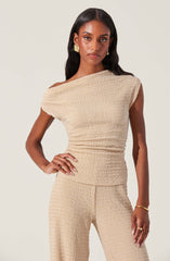 A woman is wearing a matching beige outfit consisting of a one-shoulder top with a textured, crinkled fabric and high-waisted pants made of the same textured material. The top has asymmetrical shoulder coverage, exposing one shoulder, and the pants are fitted at the waist and hips.
