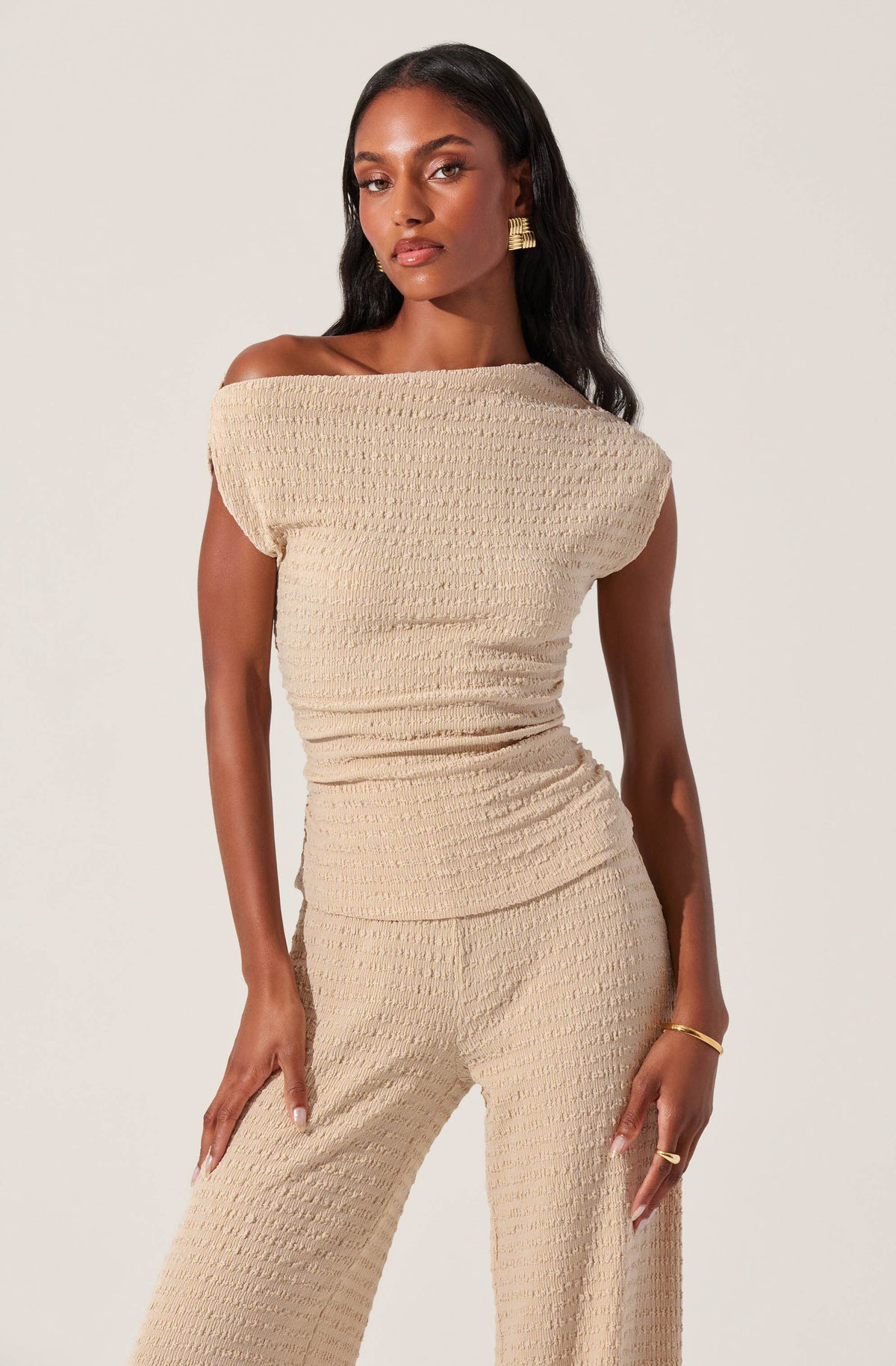 A woman is wearing a matching beige textured outfit consisting of an off-the-shoulder top with a loose fit and wide sleeves, paired with wide-leg pants made from the same fabric. The fabric has a crinkled, waffle-like texture. She accessorizes with gold earrings, a gold bracelet, and a gold ring.