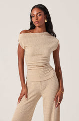 A woman is wearing a matching beige textured outfit consisting of an off-the-shoulder top with a loose fit and wide sleeves, paired with wide-leg pants made from the same fabric. The fabric has a crinkled, waffle-like texture. She accessorizes with gold earrings, a gold bracelet, and a gold ring.