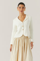 A woman is wearing an elegant white blazer with a notched lapel collar and large pearl-like buttons down the front. The blazer has a slightly textured fabric and a tailored fit. She is also wearing a flowing, high-waisted, beige skirt with gathered fabric, creating a voluminous and soft silhouette. She accessorized with gold earrings and a ring.