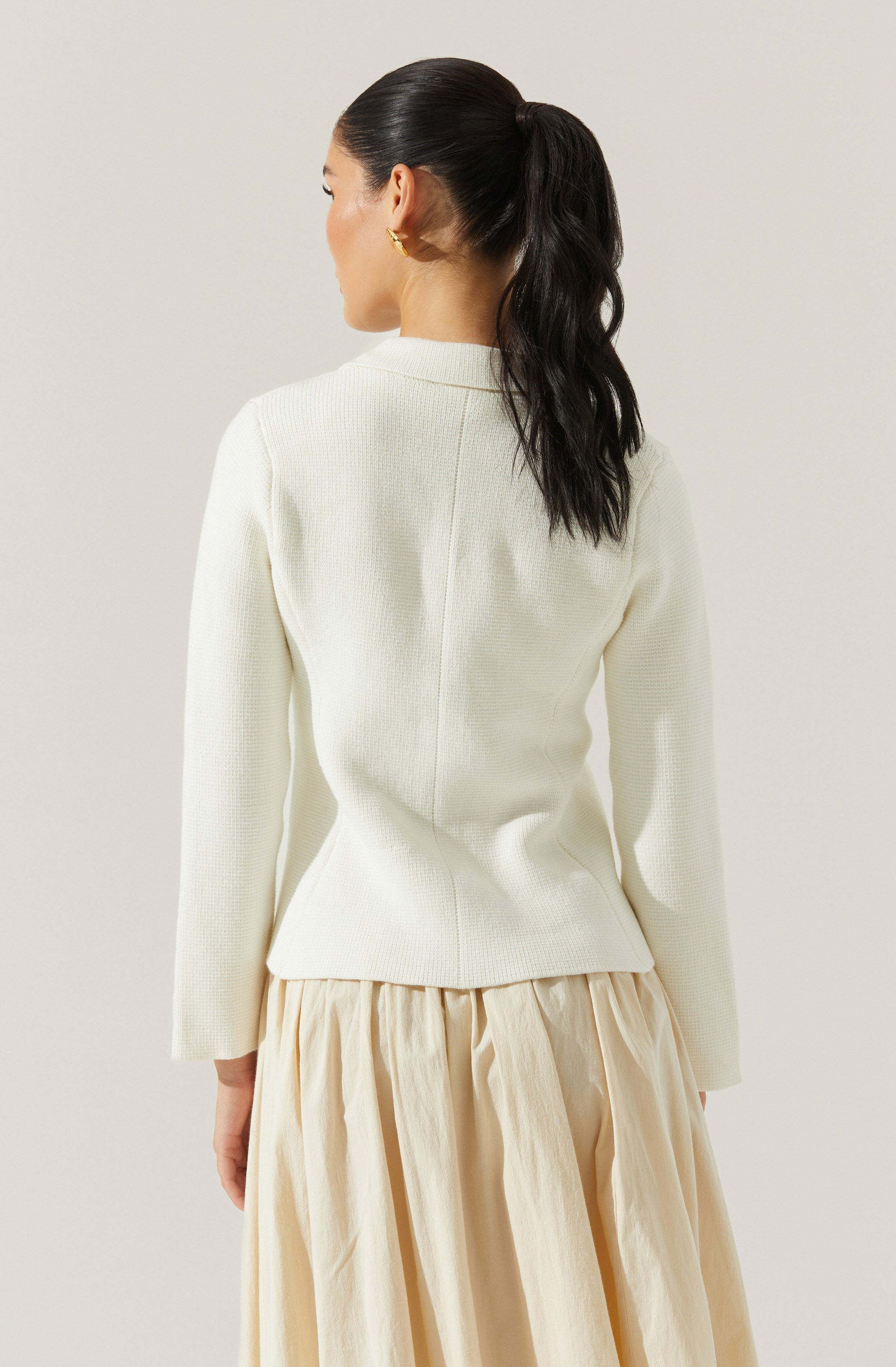 A woman is wearing a cream-colored, long-sleeved, textured sweater with a smooth finish on the collar and front. She is also dressed in a beige, pleated, ankle-length skirt made of lightweight fabric.