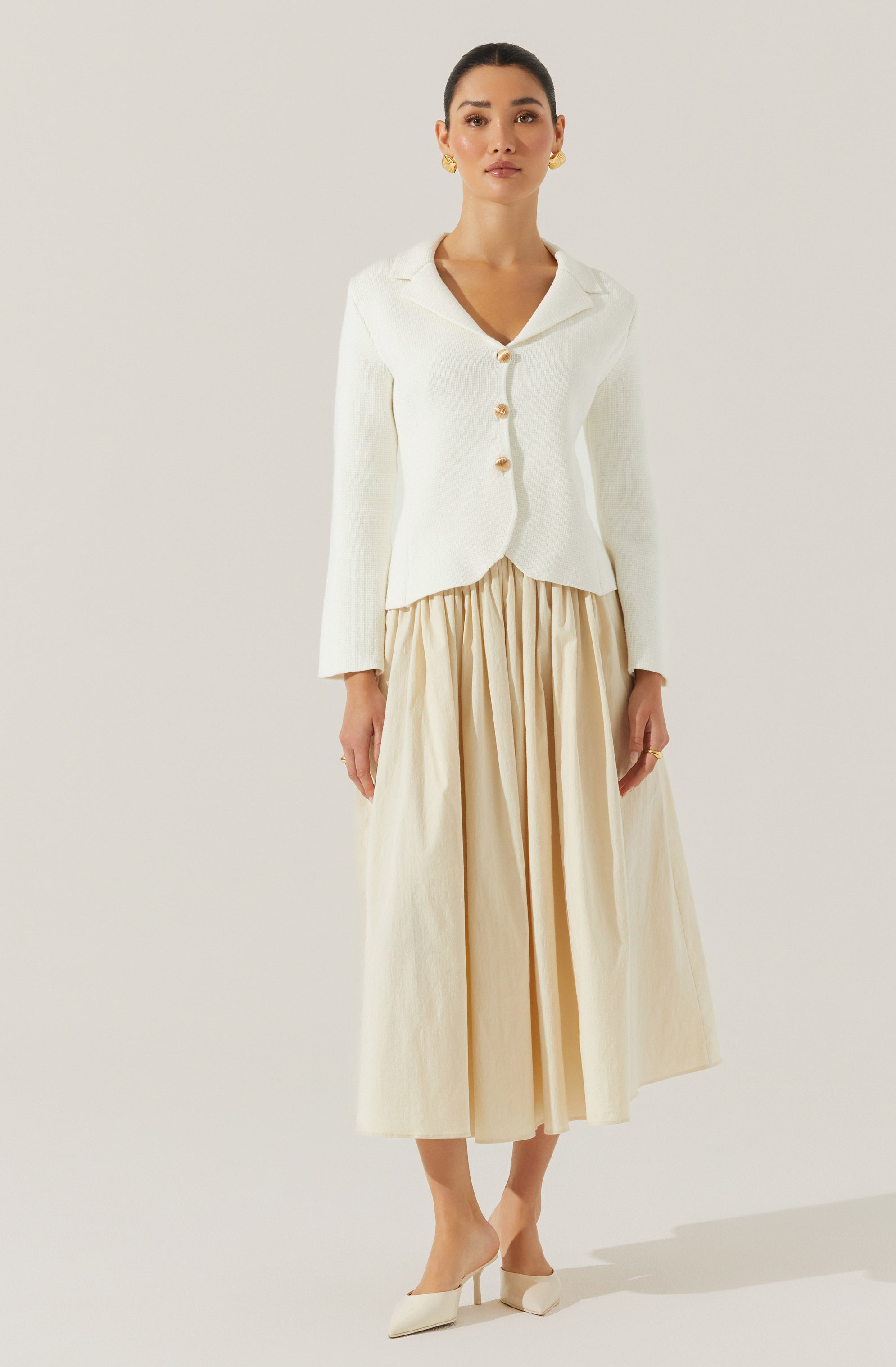 A woman is wearing a white, long-sleeved, button-up shirt with a collar. The shirt has four large, round, shiny buttons down the front. She pairs it with a high-waisted, beige, pleated, ankle-length skirt. She accessorizes with gold hoop earrings and a gold ring, and she is wearing off-white pointed-toe heels.