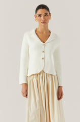 A woman is wearing a white, long-sleeved, button-up top with a collar. The top has a textured fabric and features large, round, gold buttons down the front. She pairs it with a flowing, beige, pleated skirt.