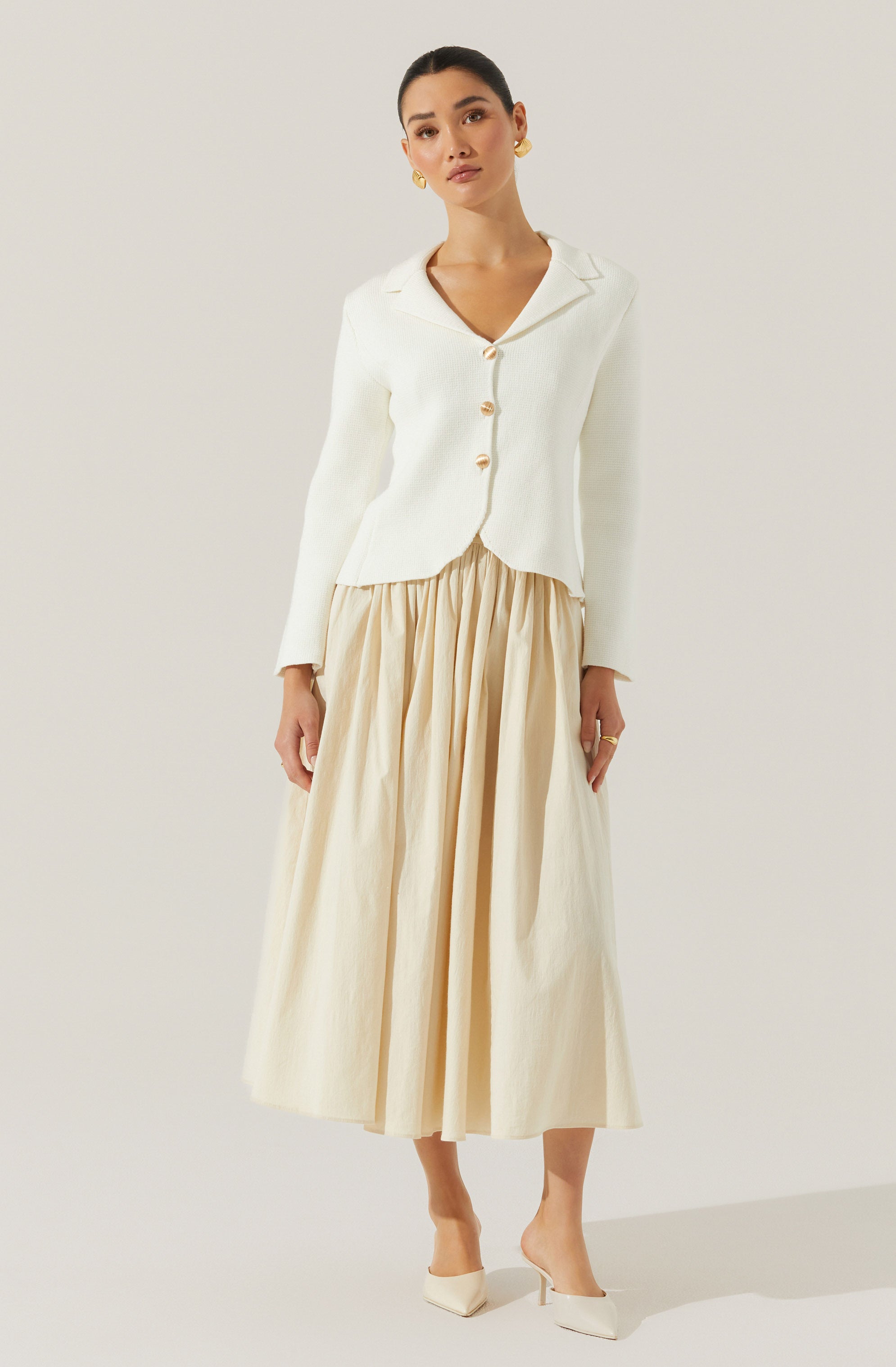 A woman is wearing a cream-colored, long-sleeved blazer with a notched collar and four large buttons down the front. The blazer has a slightly cropped fit with a pointed hem. She pairs it with a high-waisted, full-length, beige pleated skirt that reaches her ankles. She is also wearing cream-colored pointed-toe heels.
