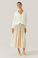 A woman is wearing a cream-colored, long-sleeved blazer with a notched collar and four large buttons down the front. The blazer has a slightly cropped fit with a pointed hem. She pairs it with a high-waisted, full-length, beige pleated skirt that reaches her ankles. She is also wearing cream-colored pointed-toe heels.