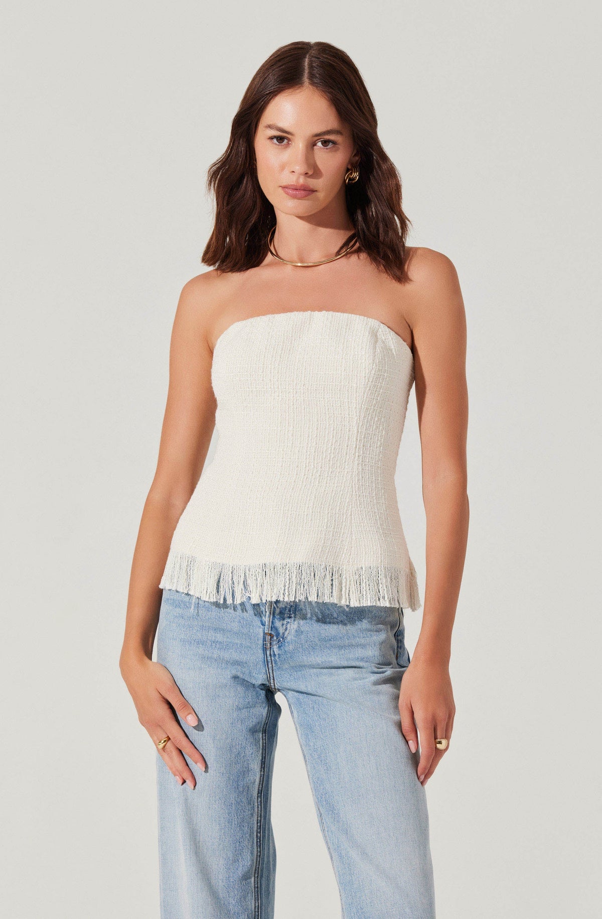 A woman is wearing a strapless, off-white, textured top with a frayed hem, paired with light-wash jeans.