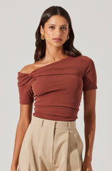 A woman is wearing a fitted, off-the-shoulder top in a warm terracotta color with short sleeves and a ruched detail across the chest. She pairs it with high-waisted, beige pleated trousers.