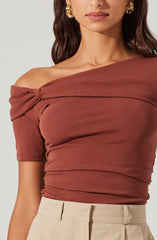 A woman is wearing an off-the-shoulder top in a warm, earthy rust color. The top features a gathered, twisted fabric detail at the shoulder, creating a slightly ruched appearance across the bust and sleeves. The short sleeves end above the elbow, and the fabric appears soft and stretchy. The top is paired with light beige high-waisted pants.