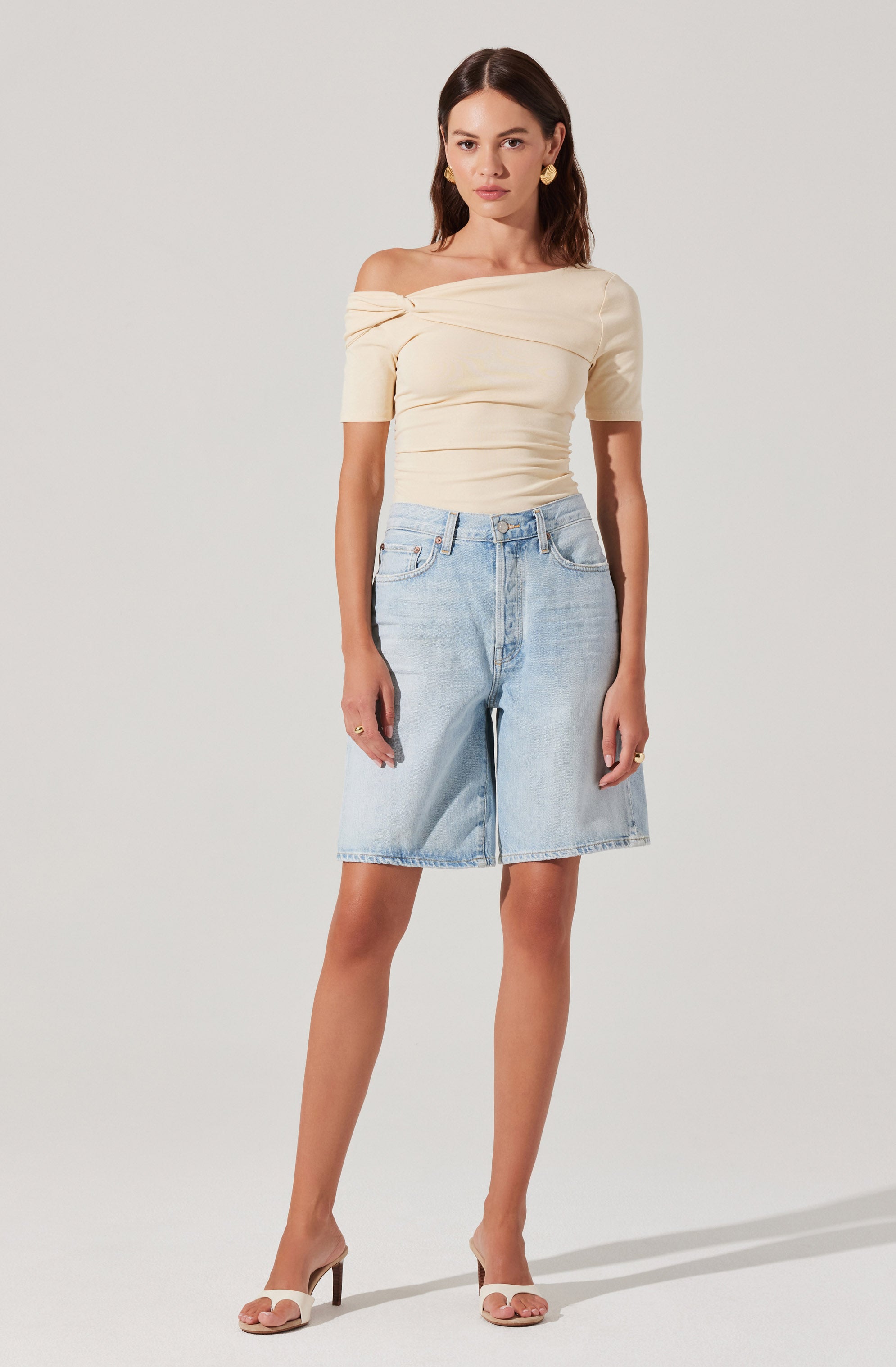 A woman is wearing an off-the-shoulder, short-sleeved, cream-colored top with gathered fabric details. She pairs it with light-wash, knee-length denim shorts that have a relaxed fit. She completes her outfit with beige high-heeled sandals and gold earrings.