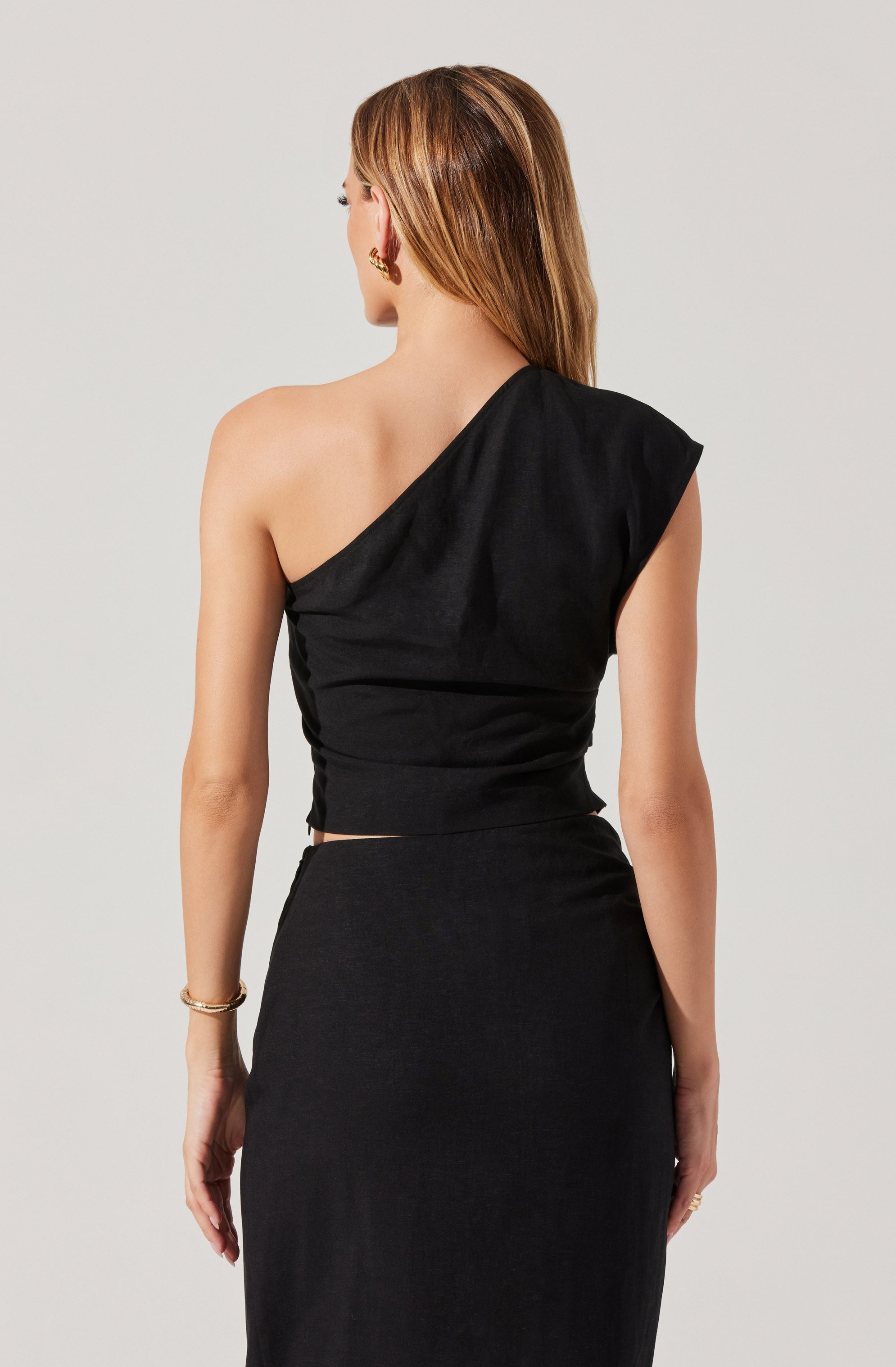 A woman is wearing a black, one-shoulder top with a draped, asymmetrical design. The top is fitted around her waist and has a slightly ruched appearance. She pairs it with a high-waisted black skirt that extends below the waist. The overall outfit is elegant and monochromatic.