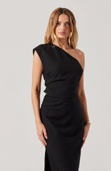 A woman is wearing a black dress with asymmetrical design details. The dress has one shoulder strap on the left side, while the right side is sleeveless with a draped, slightly ruffled fabric. The waist is fitted, and the skirt is long, straight, and reaches the floor.