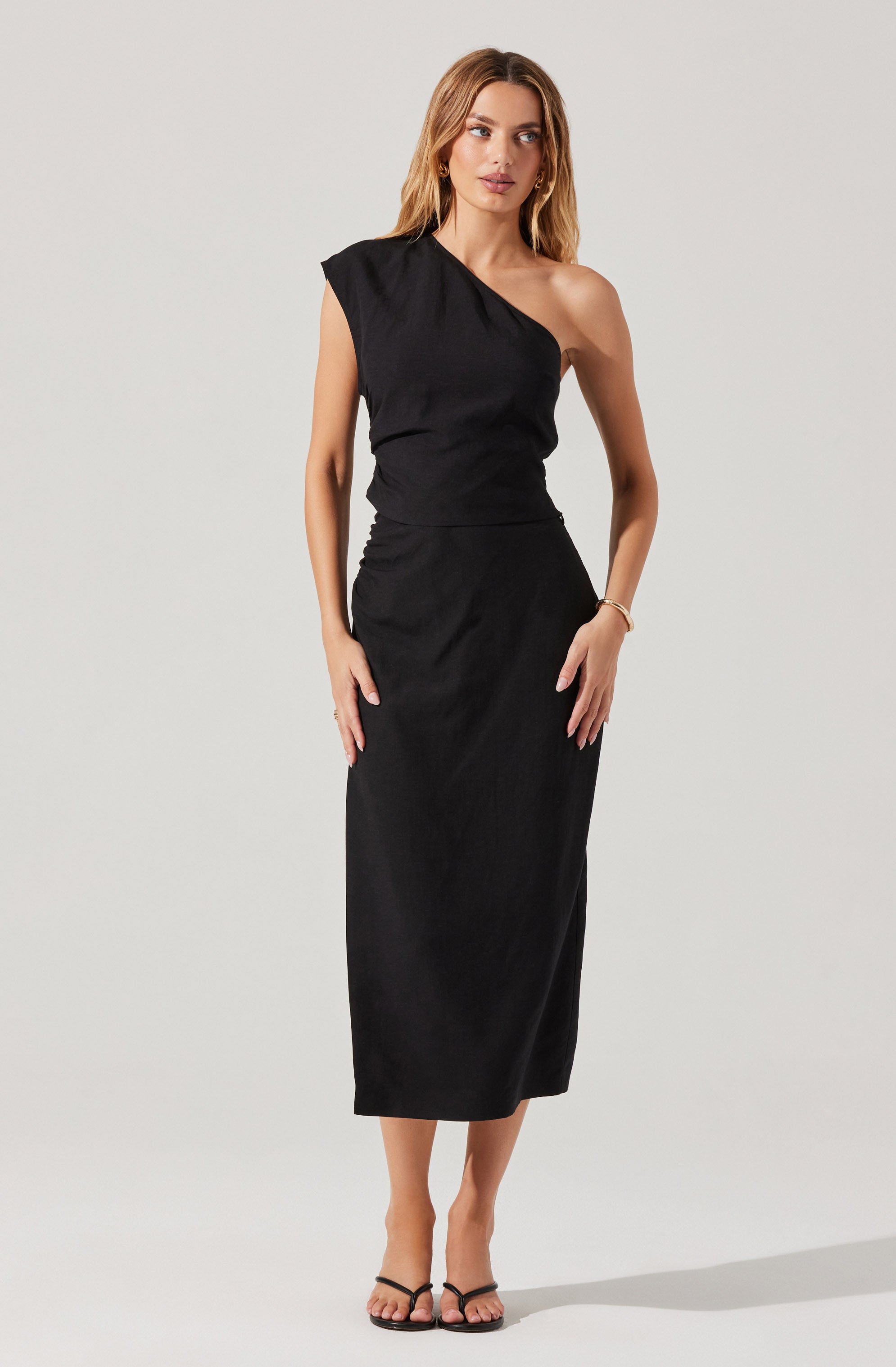 A woman is wearing a black dress with a one-shoulder design. The dress has a fitted waist and a long, straight skirt that reaches mid-calf. The fabric appears to be smooth and slightly shiny. She is also wearing black strappy heels and a gold bracelet on her right wrist.