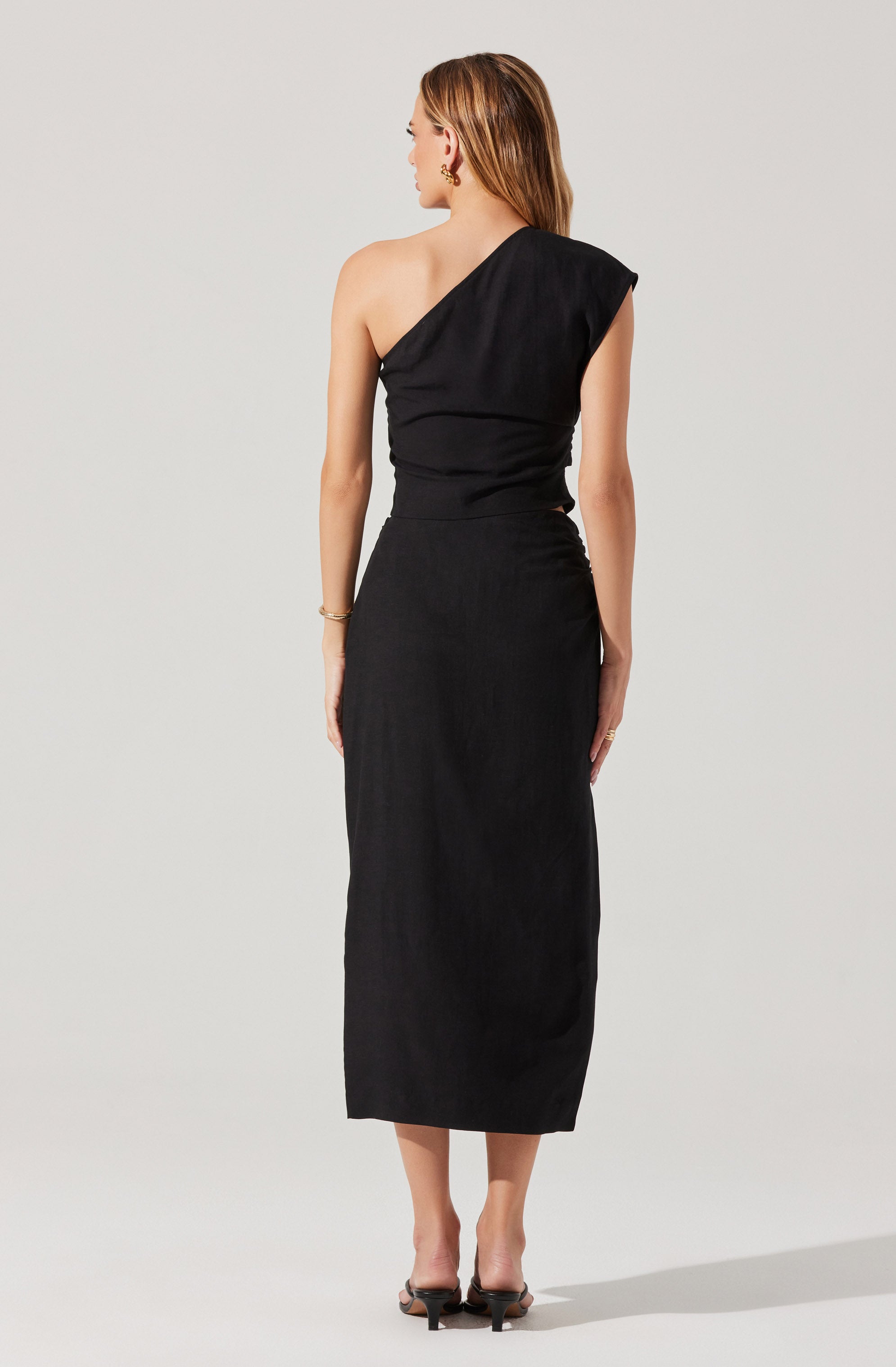 A woman is wearing a black, one-shoulder, sleeveless maxi dress that is fitted at the waist and flows down to her ankles. She has her hair styled straight and is accessorizing with gold earrings, a gold bracelet, and rings. She is standing with her back to the camera against a plain light gray background.