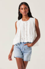 A woman is wearing a sleeveless, white embroidered top with eyelet details, scalloped edges, and a row of buttons down the front. She pairs the top with light-wash denim shorts.