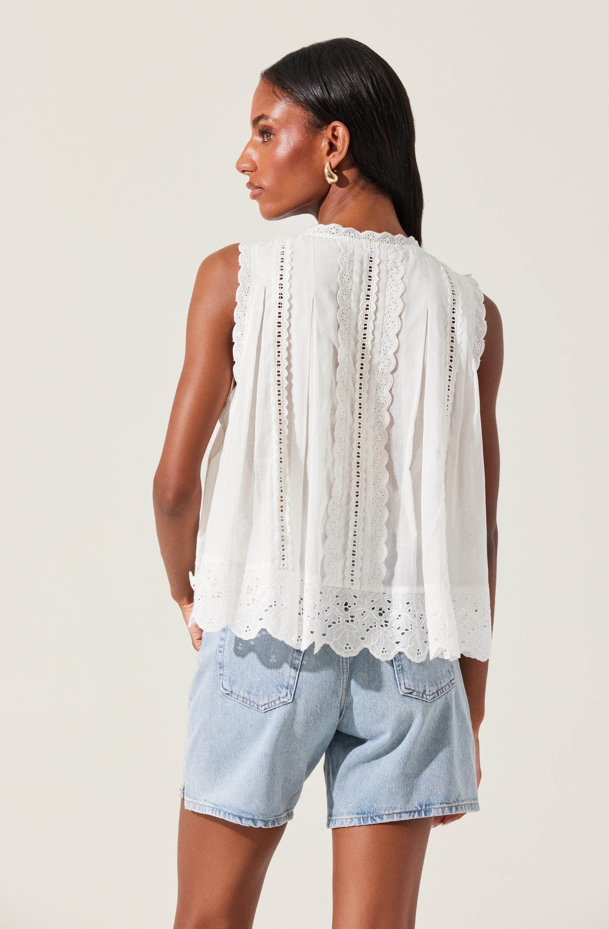 A woman is wearing a sleeveless white top with intricate eyelet and lace embroidery, featuring scalloped edges at the hem. She has paired it with light-wash denim shorts.