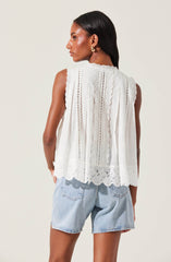 A woman is wearing a sleeveless white top with intricate eyelet and lace embroidery, featuring scalloped edges at the hem. She has paired it with light-wash denim shorts.