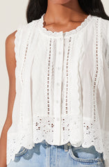 A sleeveless white blouse featuring intricate eyelet embroidery and scalloped lace trim around the neckline and armholes. The front has a vertical row of small buttons, and the fabric displays decorative cut-out patterns creating a textured, feminine look. The blouse is paired with light-wash denim jeans.