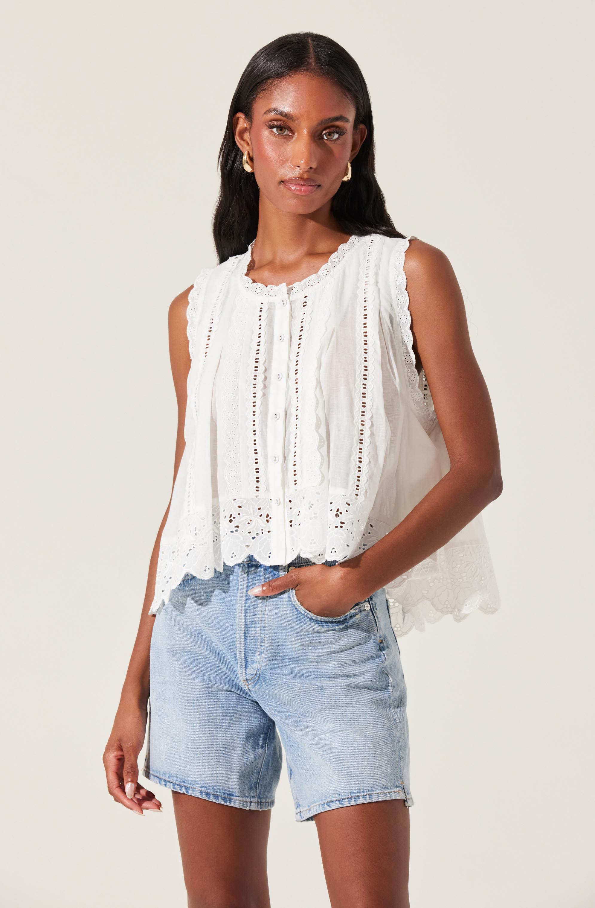 The woman is wearing a sleeveless white top with embroidered and eyelet details, featuring a scalloped hem. She pairs it with light wash denim shorts.