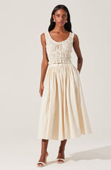 A woman is wearing a sleeveless, textured, light-colored crop top with a drawstring front and elastic waistband. She pairs it with a high-waisted, full, cream-colored midi skirt with pleats. She completes her look with open-toe heels.