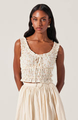 A woman is wearing a sleeveless, cropped top with ruffled shoulder straps, a gathered elastic waistband, and a front tie detail. The top features a light, pastel floral pattern. She pairs it with a high-waisted, cream-colored skirt that has a full, pleated silhouette.
