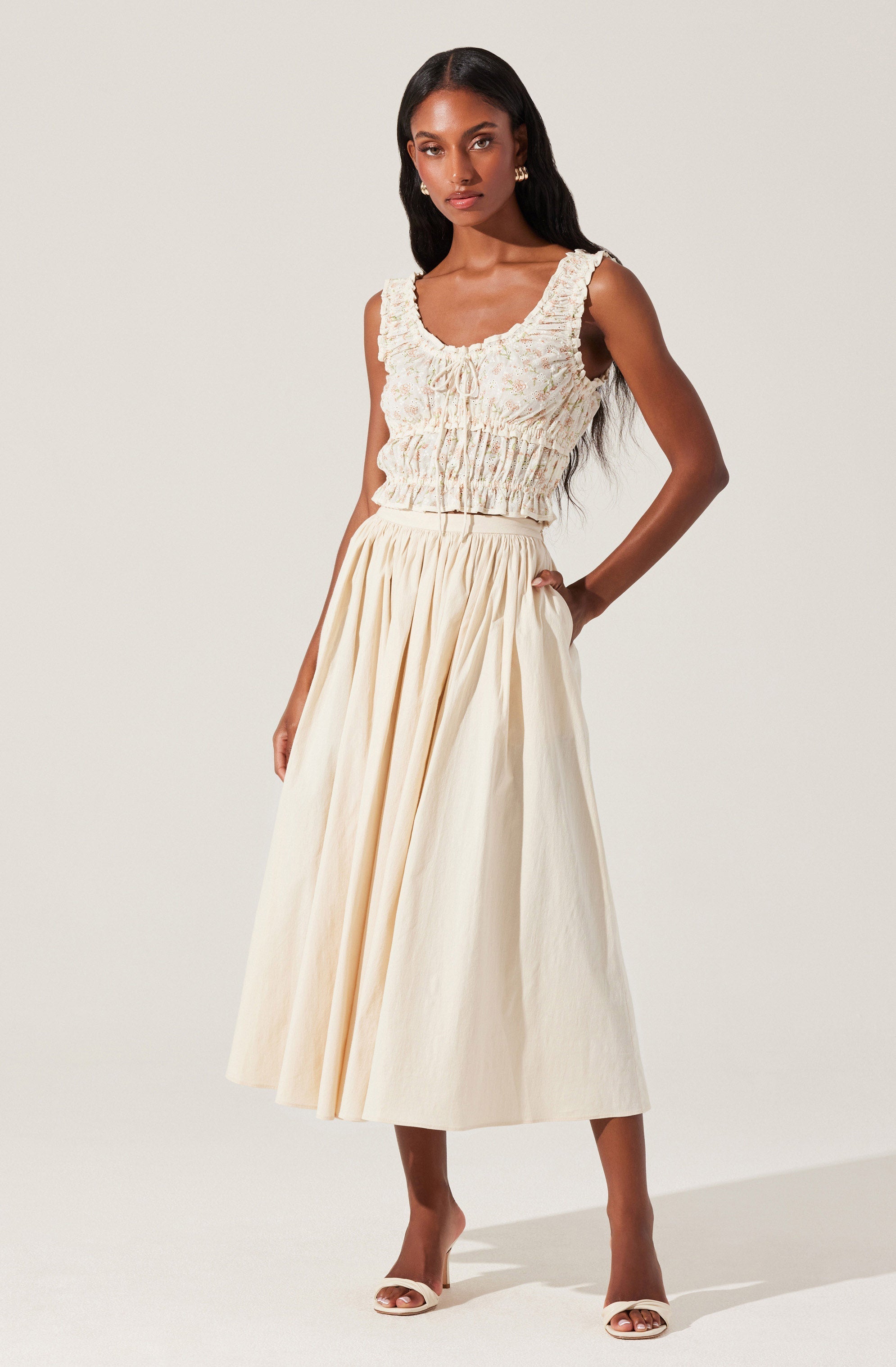 A woman is wearing a sleeveless, textured top with ruffled edges around the neckline and armholes, featuring a gathered and slightly cropped fit. She pairs it with a high-waisted, full-length, cream-colored skirt that has pleats, creating a flowing silhouette. She is wearing open-toed, heeled sandals.