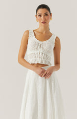 A woman is wearing a sleeveless white eyelet crop top with ruffled edges around the armholes and a gathered, elasticized waist. She is also wearing a matching white long skirt with an eyelet pattern and a high waist. She accessorizes with gold earrings and multiple rings.