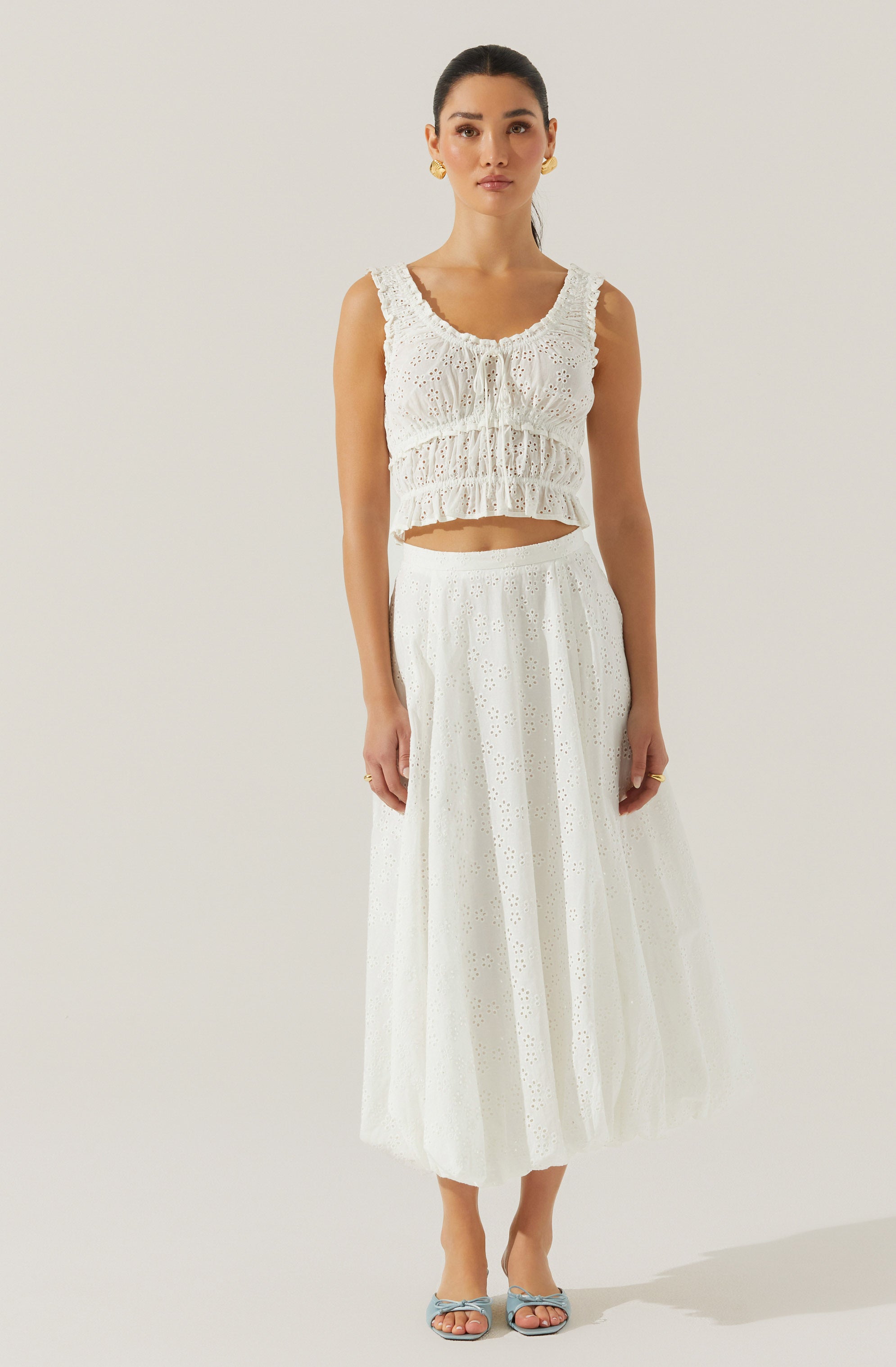 A woman is wearing a sleeveless, white, eyelet crop top with ruffled details and a scoop neckline. She pairs it with a high-waisted, white, eyelet maxi skirt that features a subtle pattern of small holes and a flowy silhouette. She completes her outfit with light-colored open-toe heels and gold earrings.
