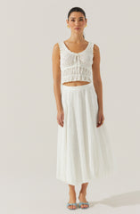 A woman is wearing a sleeveless, white, eyelet crop top with ruffled details and a scoop neckline. She pairs it with a high-waisted, white, eyelet maxi skirt that features a subtle pattern of small holes and a flowy silhouette. She completes her outfit with light-colored open-toe heels and gold earrings.