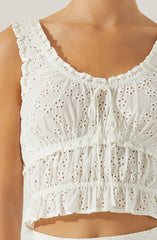 A white sleeveless crop top with eyelet embroidery featuring floral and circular patterns. The top has ruffled shoulder straps, a ruffled neckline with a tie at the center, and a gathered, elasticized hem with ruffles.