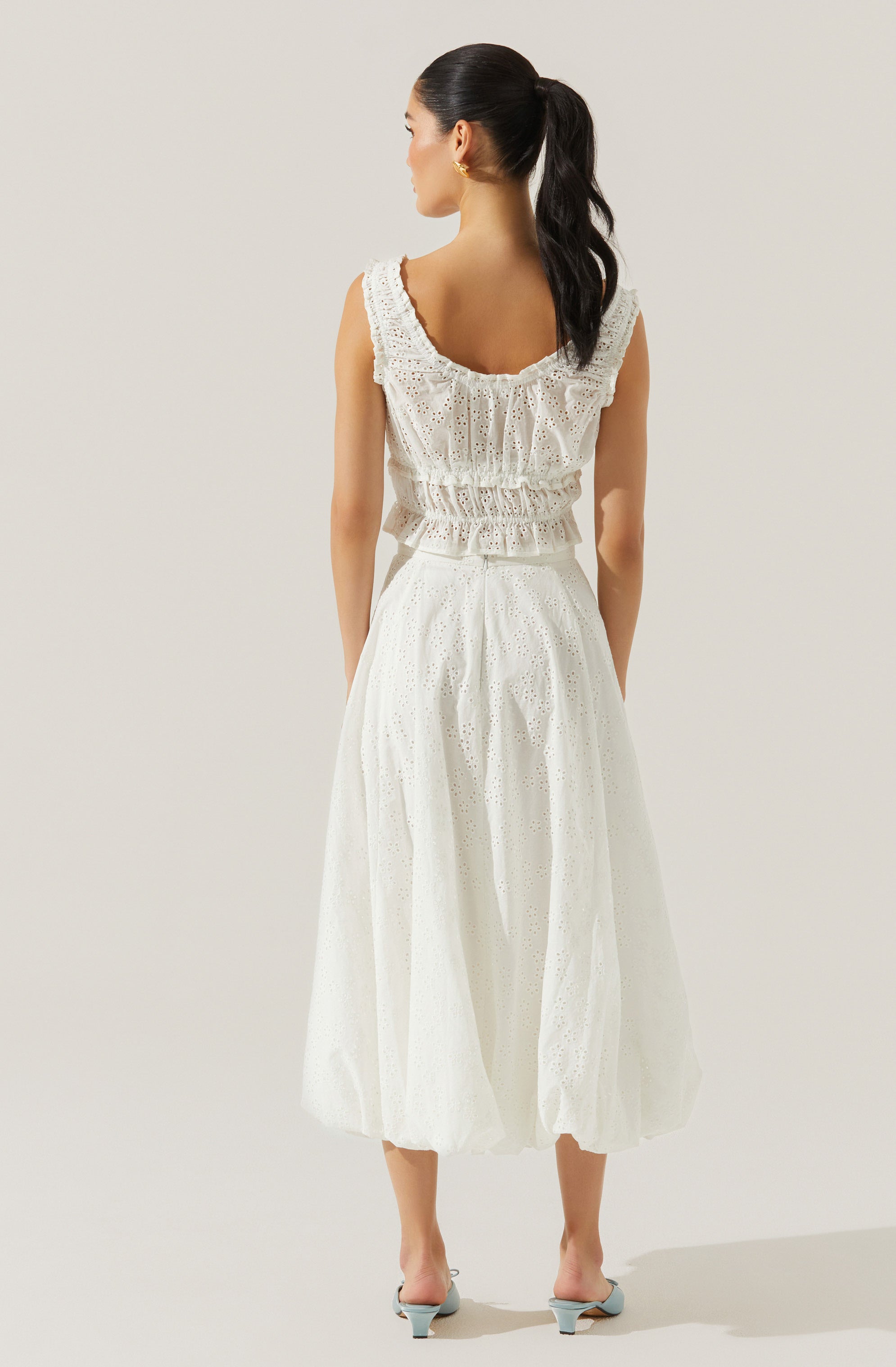 A woman is wearing a sleeveless, white eyelet dress with ruffle details on the neckline and waist. The dress has a loose, flowing skirt reaching mid-calf length. She has a high ponytail hairstyle and is wearing small hoop earrings.