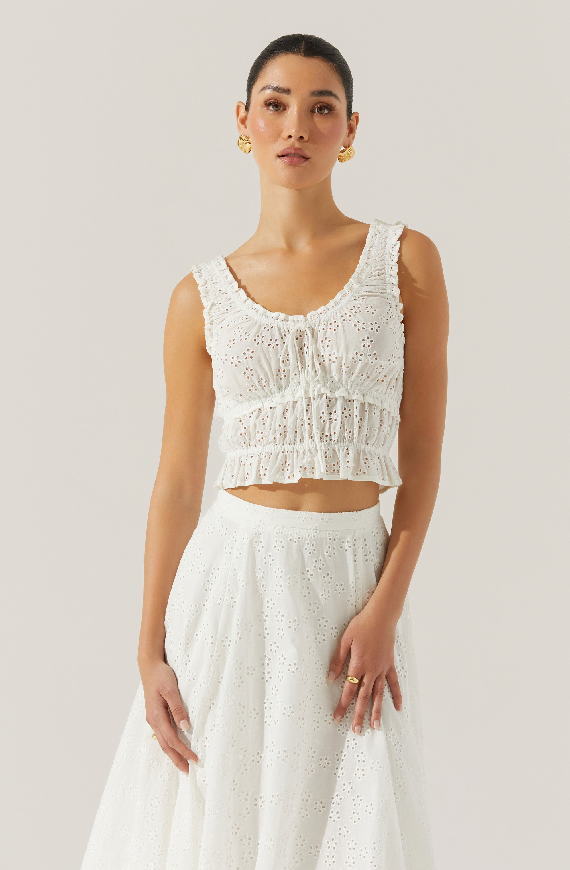 A woman is wearing a sleeveless white top with eyelet embroidery, featuring a round neckline, gathered details, and a ruffled hem. She pairs it with a matching white skirt that also has eyelet embroidery, creating a coordinated and summery look. She accessorizes with large gold earrings and a gold ring.