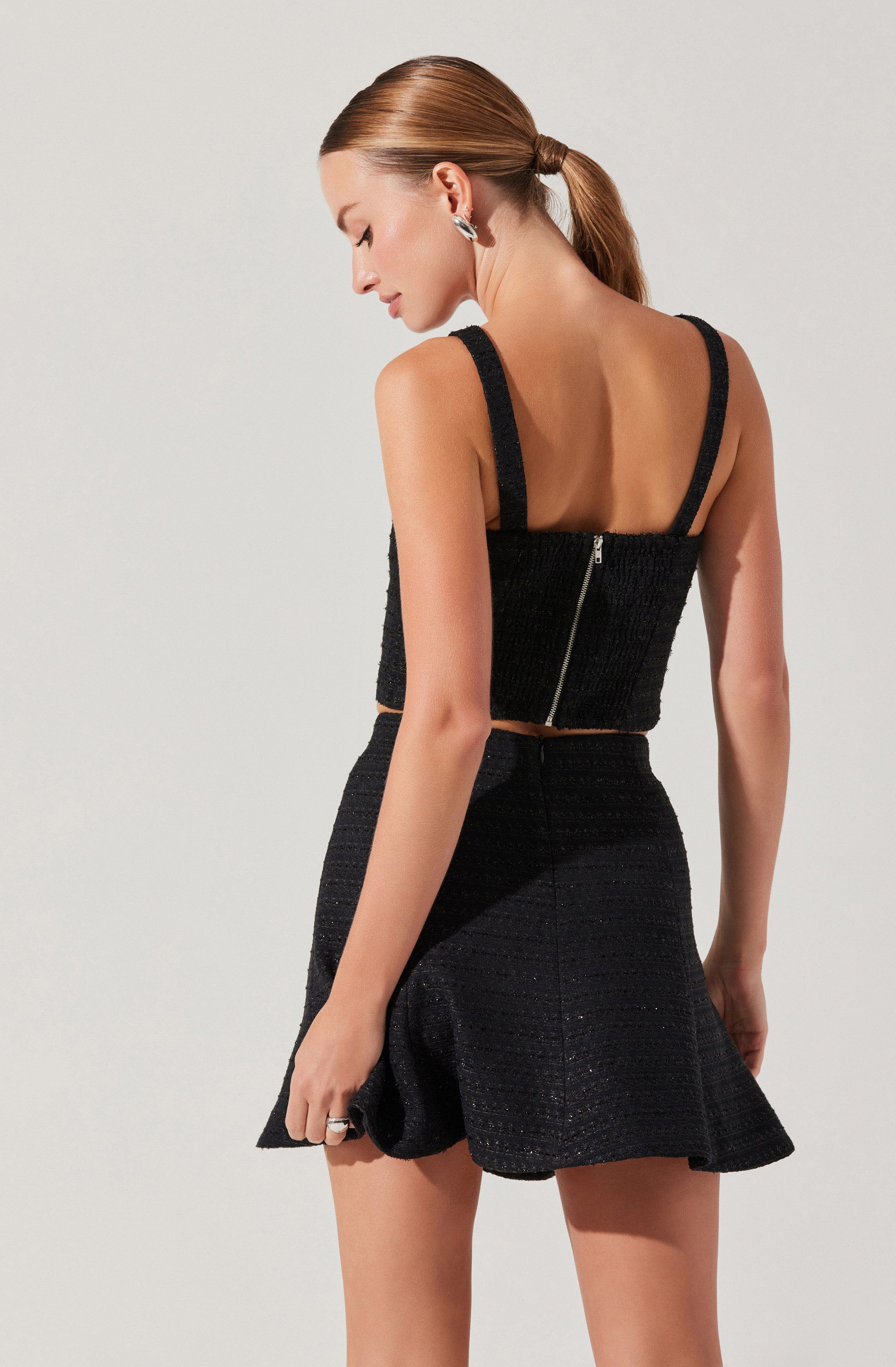 A woman is wearing a two-piece black outfit consisting of a sleeveless, fitted top with thick shoulder straps and a visible back zipper, paired with a high-waisted, flared skirt. The fabric appears textured with subtle horizontal stripes.