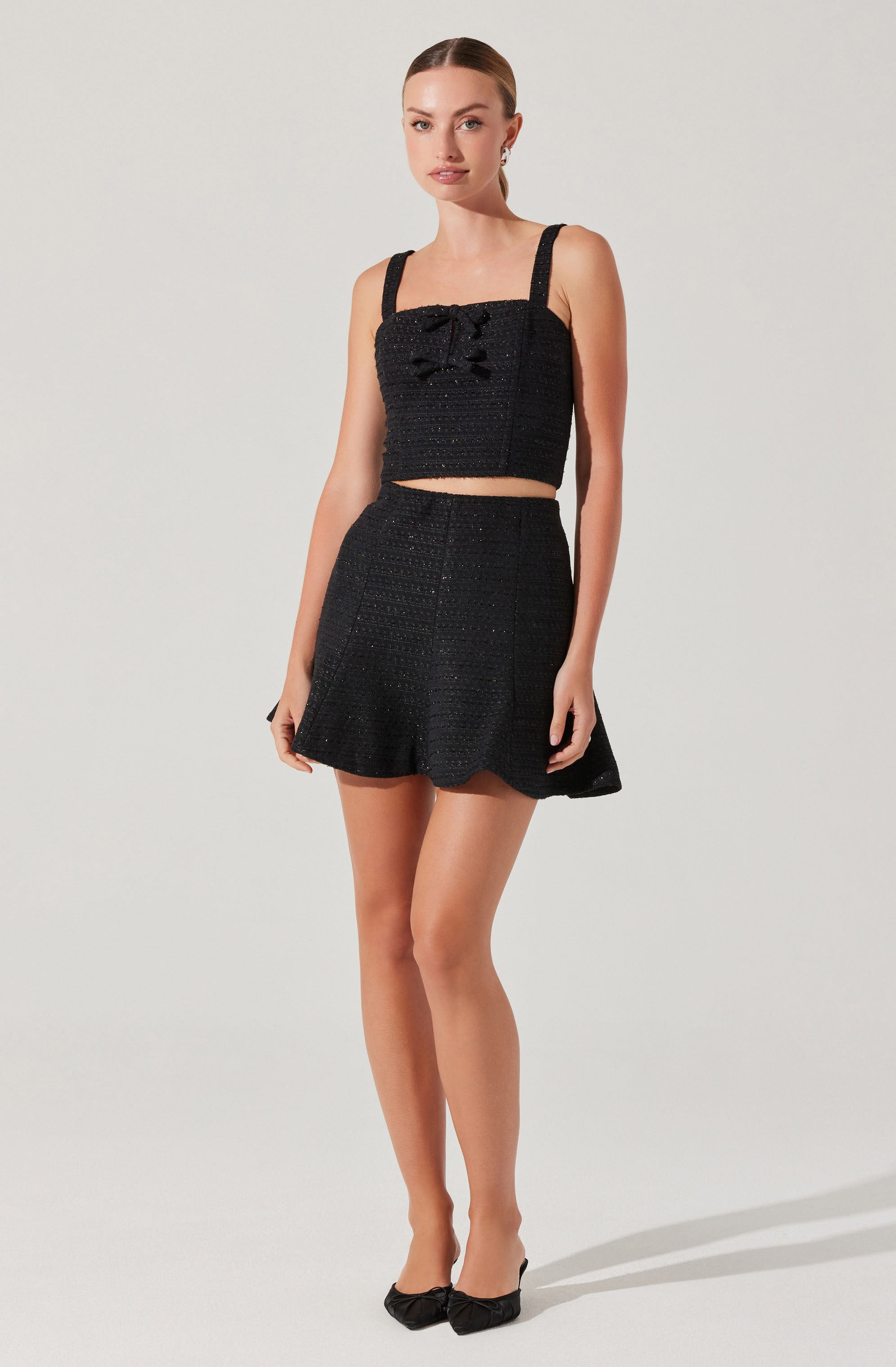 A woman is wearing a black two-piece outfit consisting of a sleeveless crop top with wide straps and a bow detail at the front, paired with a matching high-waisted skirt that has a slightly flared hem. She is also wearing black pointed-toe heels.