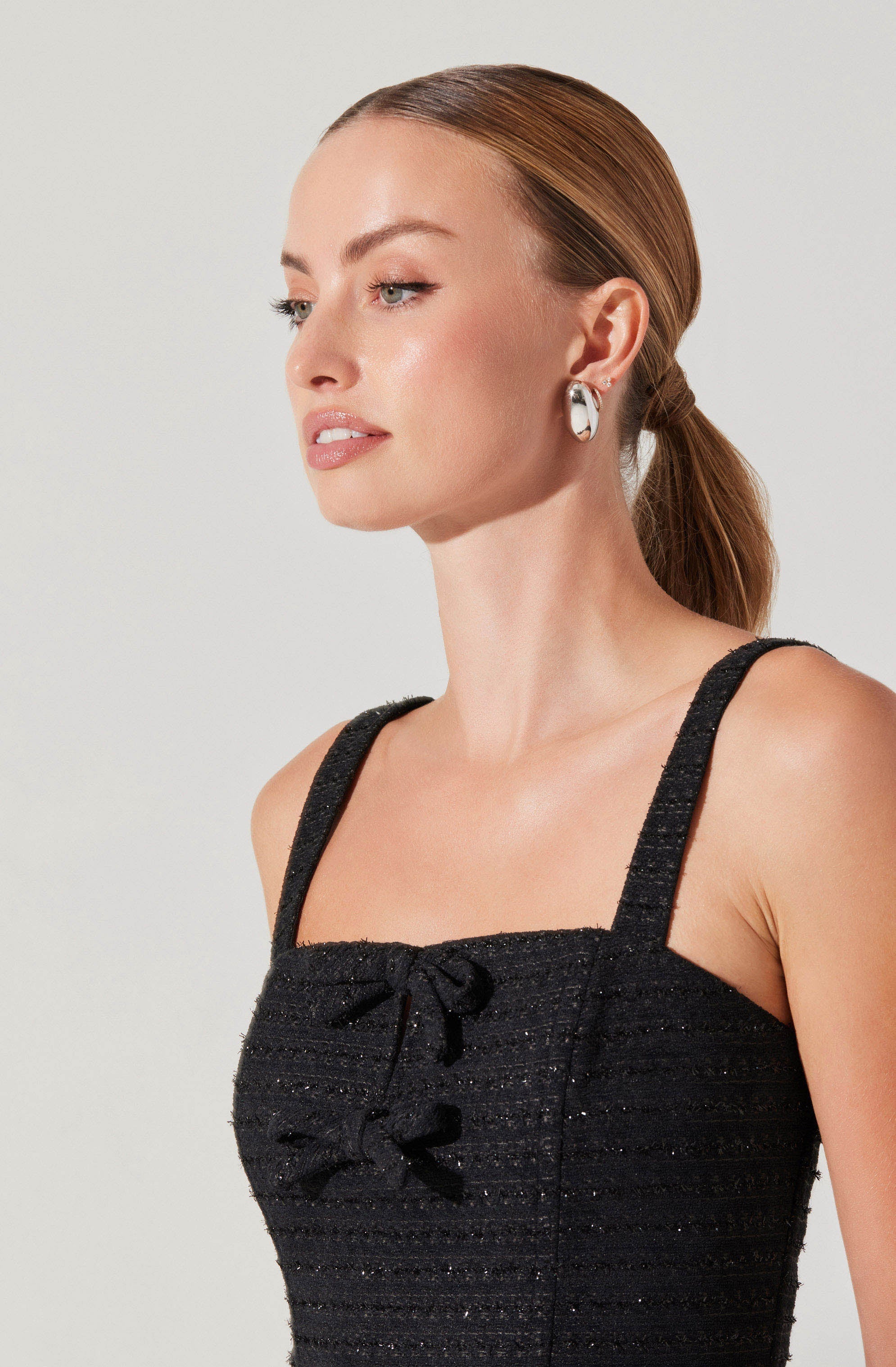 A woman is wearing a black textured sleeveless top with spaghetti straps and decorative bows on the front. She has her hair styled in a low ponytail and is accessorized with large, white and black patterned earrings.