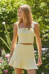 A woman is wearing a matching sleeveless top and high-waisted skirt set in a light, creamy yellow fabric. The top features two small bows on the front, and the skirt has an A-line silhouette. She is outdoors surrounded by greenery and pink flowers.