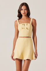 A young woman is wearing a matching yellow two-piece outfit. The top is a sleeveless, cropped design with wide shoulder straps and features a decorative bow on the front. The high-waisted skirt flares out slightly and has a textured, horizontal striped pattern.