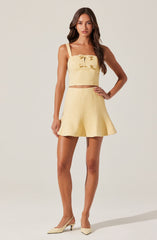 A woman is wearing a matching light yellow outfit consisting of a sleeveless crop top with thick straps and a bow at the chest, paired with high-waisted, flared shorts. She is also wearing cream-colored high heels.