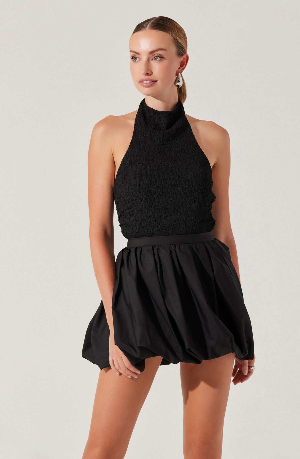 A woman wearing a sleeveless black textured halter top with a high neckline, paired with a high-waisted black skirt that has a voluminous, bubble style with gathered fabric. She has her hair pulled back and is accessorized with signature earrings.