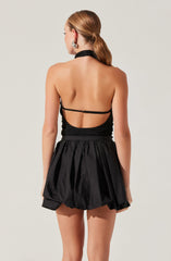 A woman with her back turned, wearing a sleeveless black top with thin straps and open back design, paired with a high-waisted, ball-shaped black skirt with a gathered, voluminous style. She has her hair styled in a low ponytail and is accessorized with large, statement earrings.