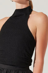 A woman is wearing a sleeveless black top with a high neckline and textured fabric. The top fits snugly and has a smooth, gathered waistband.