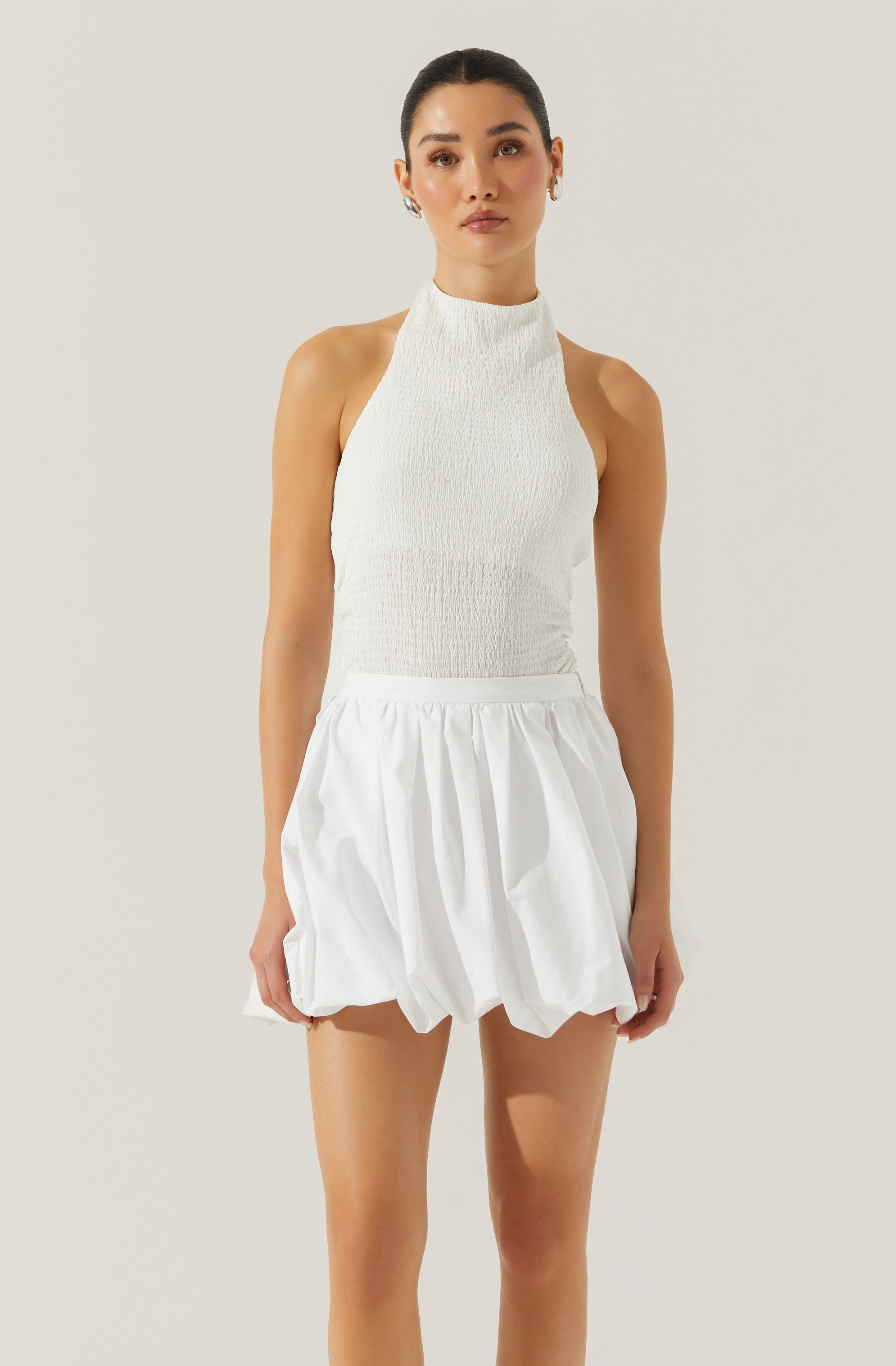 A woman is wearing a sleeveless, high-neck white top with a textured, smocked fabric and a pair of voluminous, gathered white shorts with a slightly wrinkled appearance.