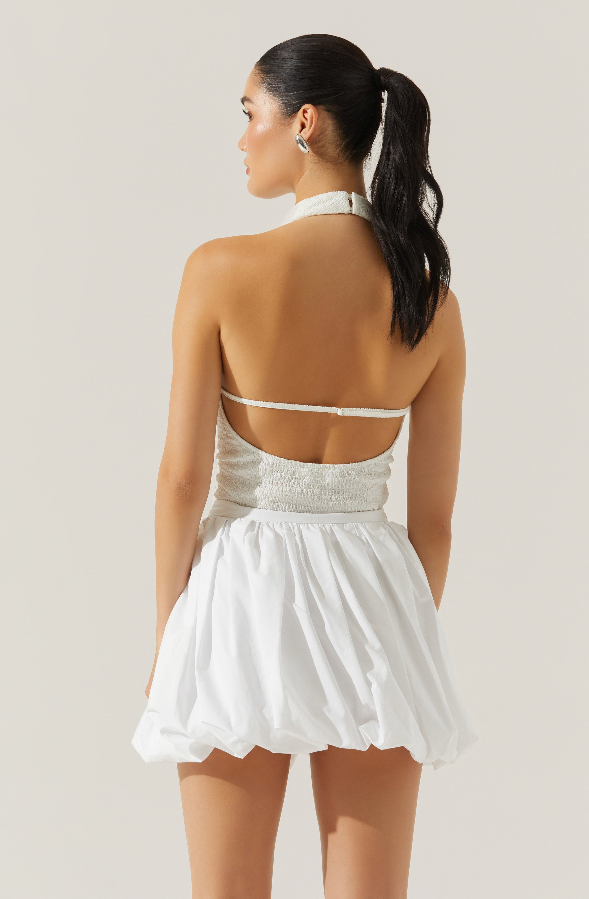 A woman is wearing a white halter top with a smocked bodice and a thin strap around her back. She pairs it with a white, puffed, knee-length skirt that has a gathered, voluminous design. Her hair is styled in a high ponytail, and she is accessorized with large, hoop earrings.