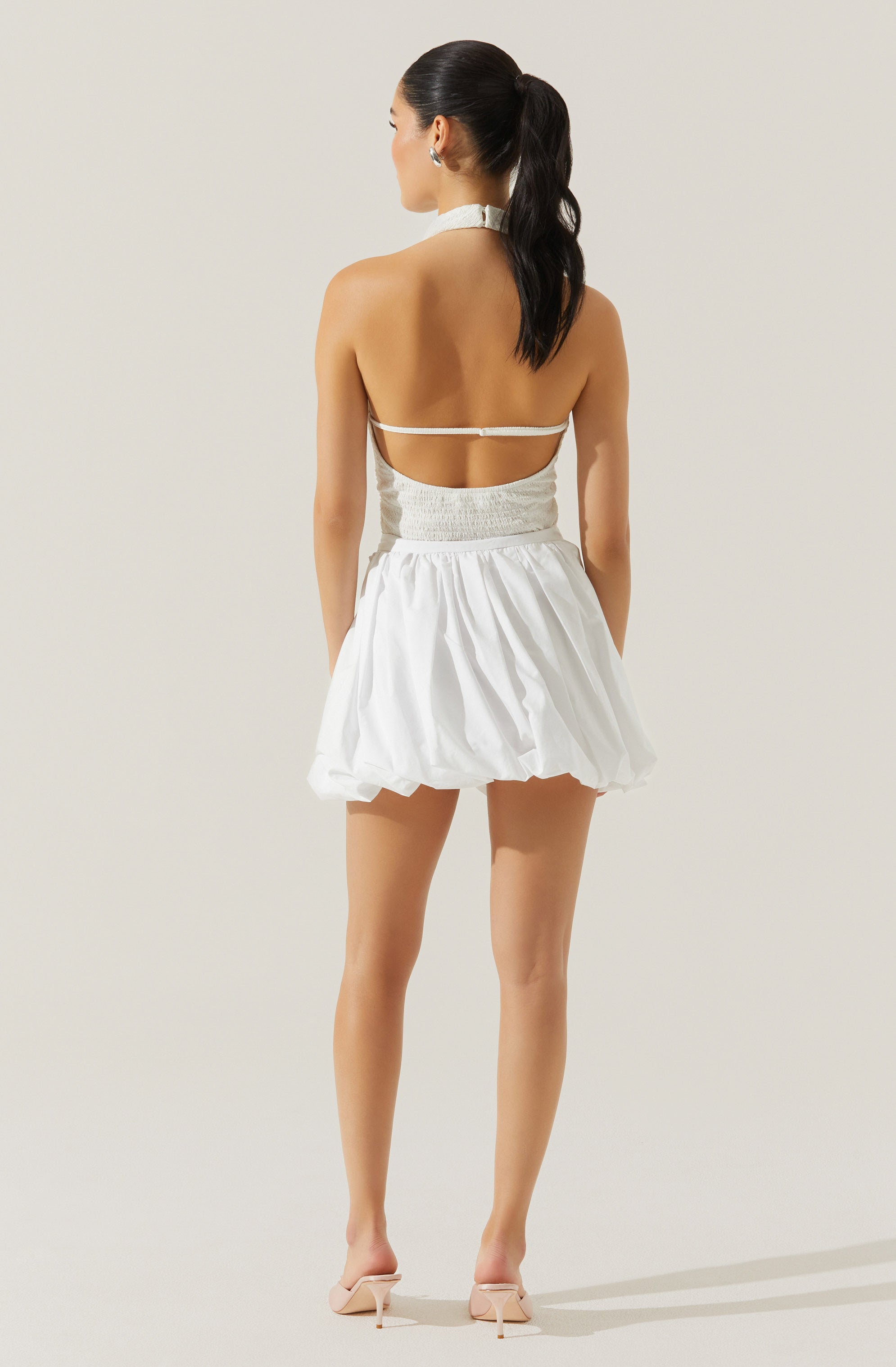 A woman is wearing a white, strapless, fitted top with a high waist and a short, puffy skirt. She has a matching white choker necklace and is wearing beige high-heeled shoes. Her long, dark hair is styled in a high ponytail.