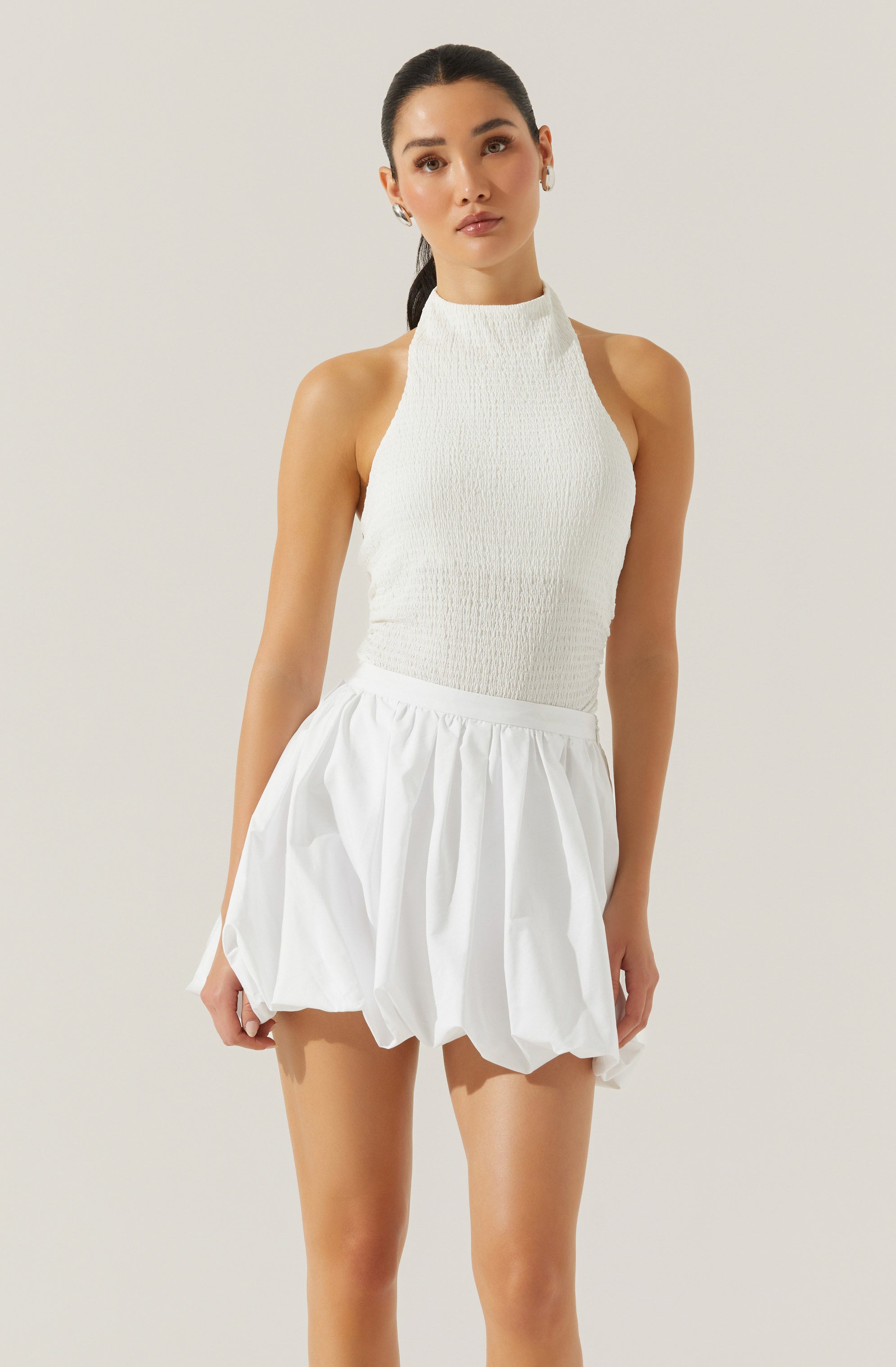 A woman is wearing a sleeveless, high-neck white top with a textured fabric and a short, white, voluminous skirt with gathered, puffed details.