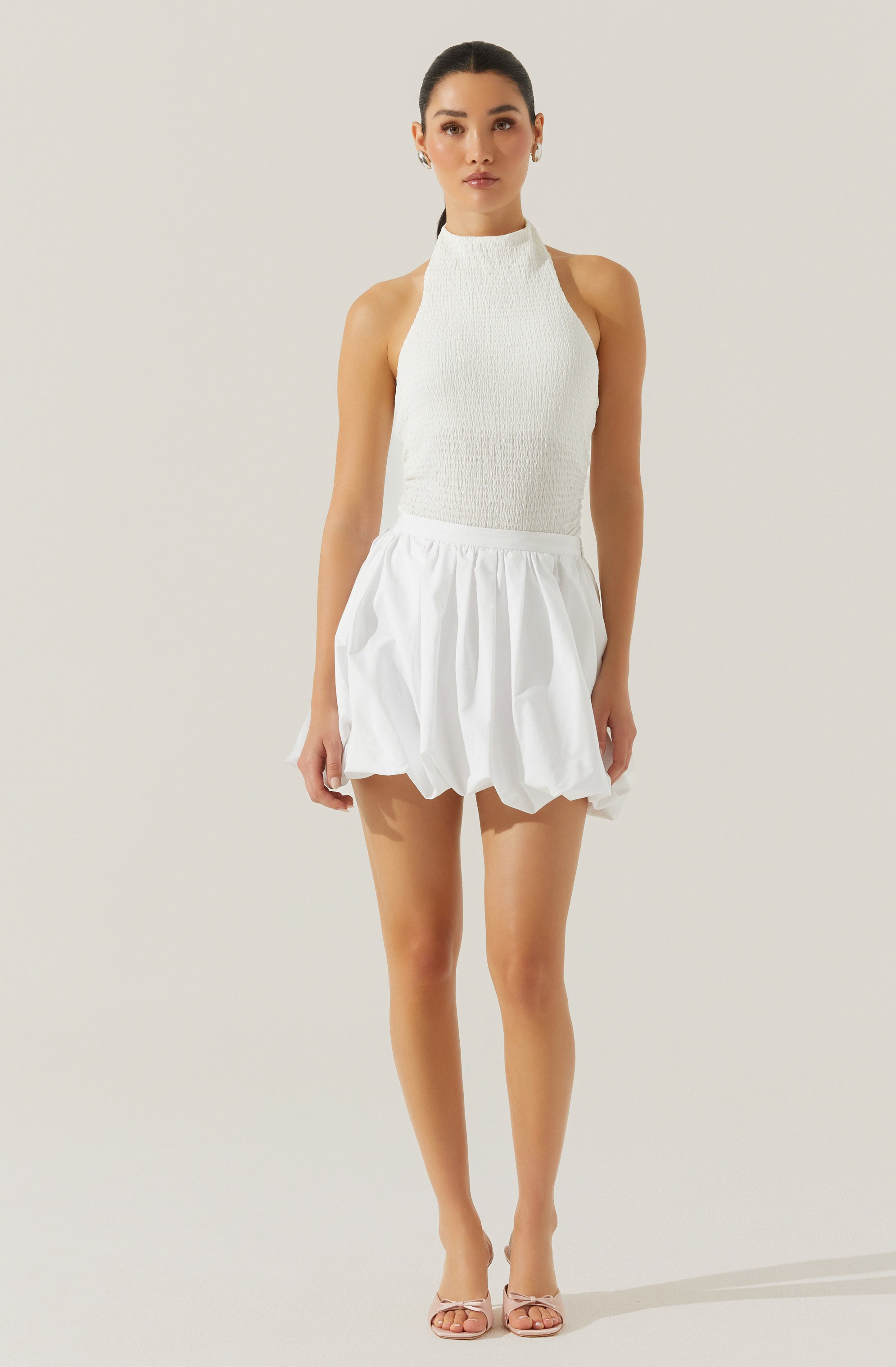 The woman is wearing a sleeveless, high-neck, textured white top paired with a white, voluminous, gathered skirt that has a bubble-like shape. She is also wearing open-toed, nude-colored heels.