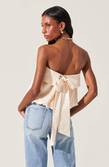 A woman is wearing a sleeveless, off-white satin top with a large bow tied at the back, creating a ruffled effect around the waist. She pairs this with light blue, high-waisted jeans. She accessorizes with gold hoop earrings and a chunky gold chain necklace.