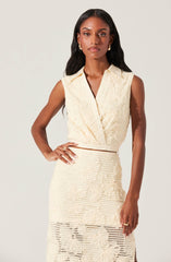 A woman is wearing a sleeveless, cream-colored dress with a textured, lace-like pattern. The dress features a deep V-neckline with a collar, a fitted waist, and a slit on the side of the skirt. She is accessorized with gold hoop earrings and rings, and has her hair styled in loose waves.