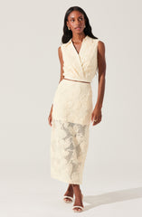 A woman is wearing a sleeveless, cream-colored knit two-piece outfit consisting of a V-neck crop top and a high-waisted, long skirt with slit and cut-out details. She is also wearing open-toe high heels.